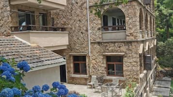 A multi-story stone building with balconies, outdoor seating, and a garden area with blue flowers.
