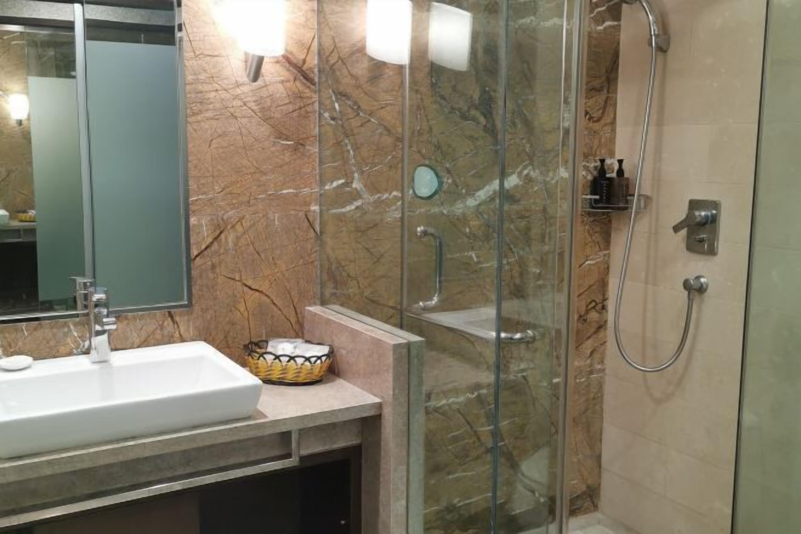 A bathroom with a marble wall, a glass shower enclosure, a sink with a soap dish, and a small basket.