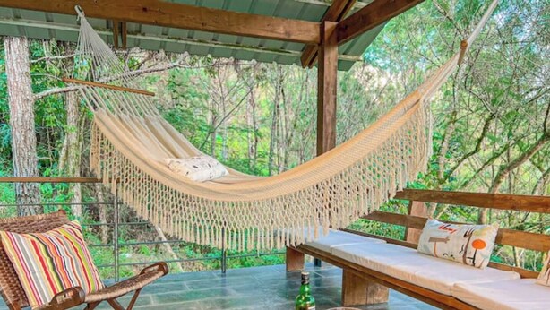 A hammock, wooden bench, and cushioned seating area with a glass table, surrounded by a forested environment.