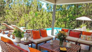 A covered outdoor seating area with wooden furniture and orange cushions, overlooking a swimming pool.