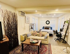 A living room with a sofa, coffee table, and a TV. There is a bed with a headboard and a desk with a chair.