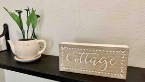 Een houten bord met de tekst 'Cottage' op een zwart aanrecht, samen met een witte theepot en een potplant.