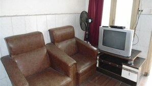 Une pièce avec deux fauteuils en cuir marron, un téléviseur posé sur un meuble en bois, un ventilateur fixé au mur et une fenêtre avec des rideaux.