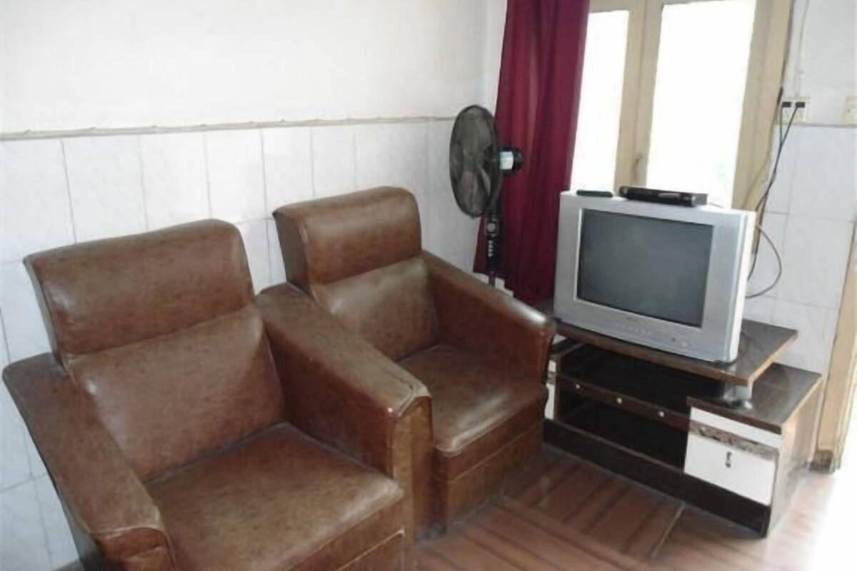 Een kamer met twee bruine leren fauteuils, een televisie op een houten standaard, een wandventilator en een raam met gordijnen.