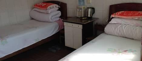 Quarto pequeno e simples com duas camas de solteiro, uma mesinha com chaleira e um bebedouro.