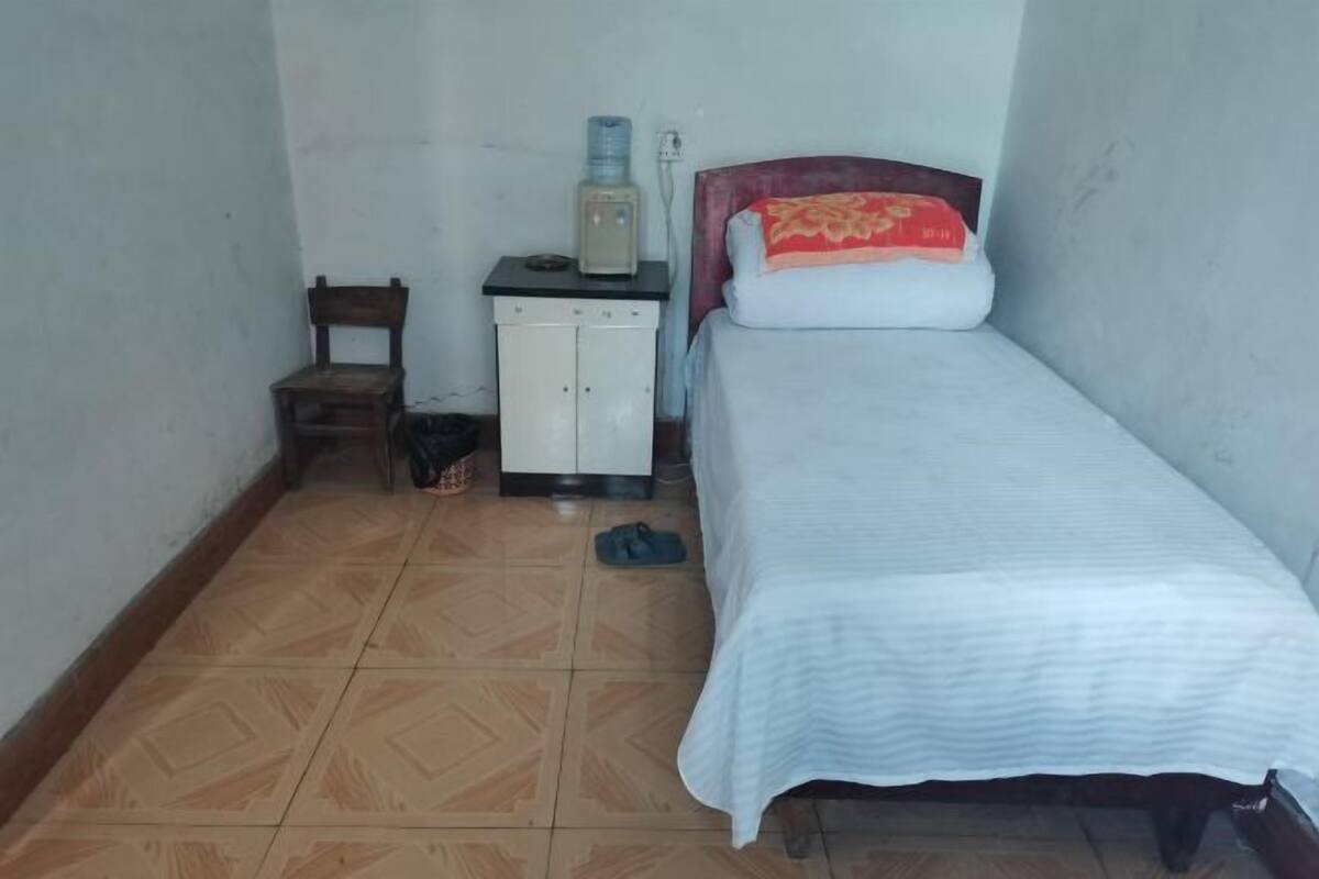 Een kleine kamer met een enkel bed, een houten stoel, een witte kast en een waterdispenser.