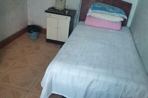 Une petite chambre avec un lit simple, un sol carrelé, une table de chevet avec une lampe et une poubelle.