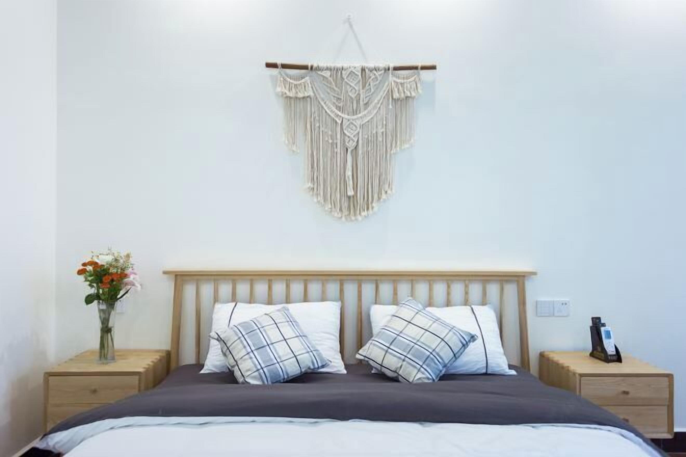A bedroom with a wooden headboard, two bedside tables, a macrame wall hanging, and a vase with flowers.