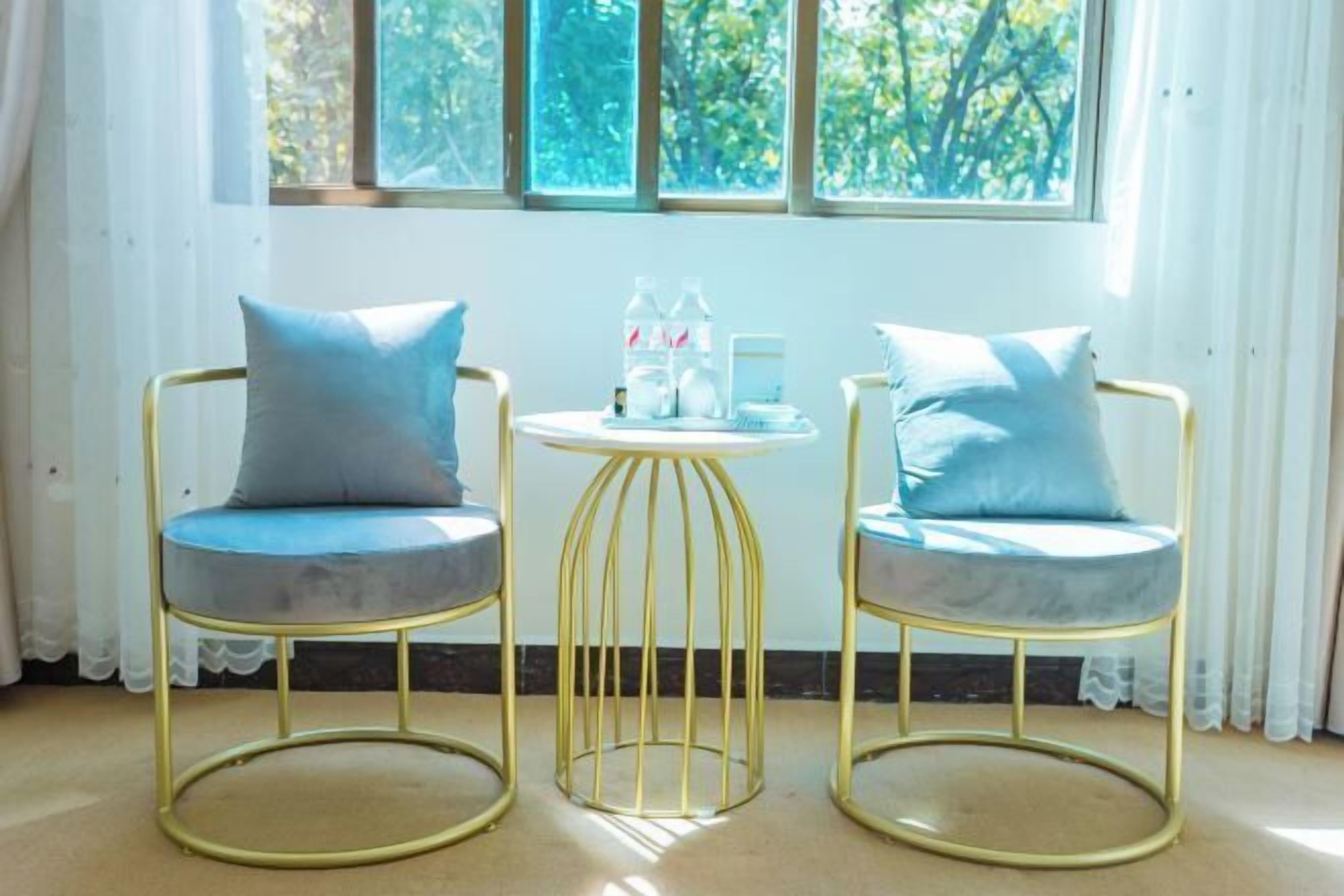 Two gold-framed chairs with blue cushions and a small round table with a white base, positioned in front of a window with white curtains.