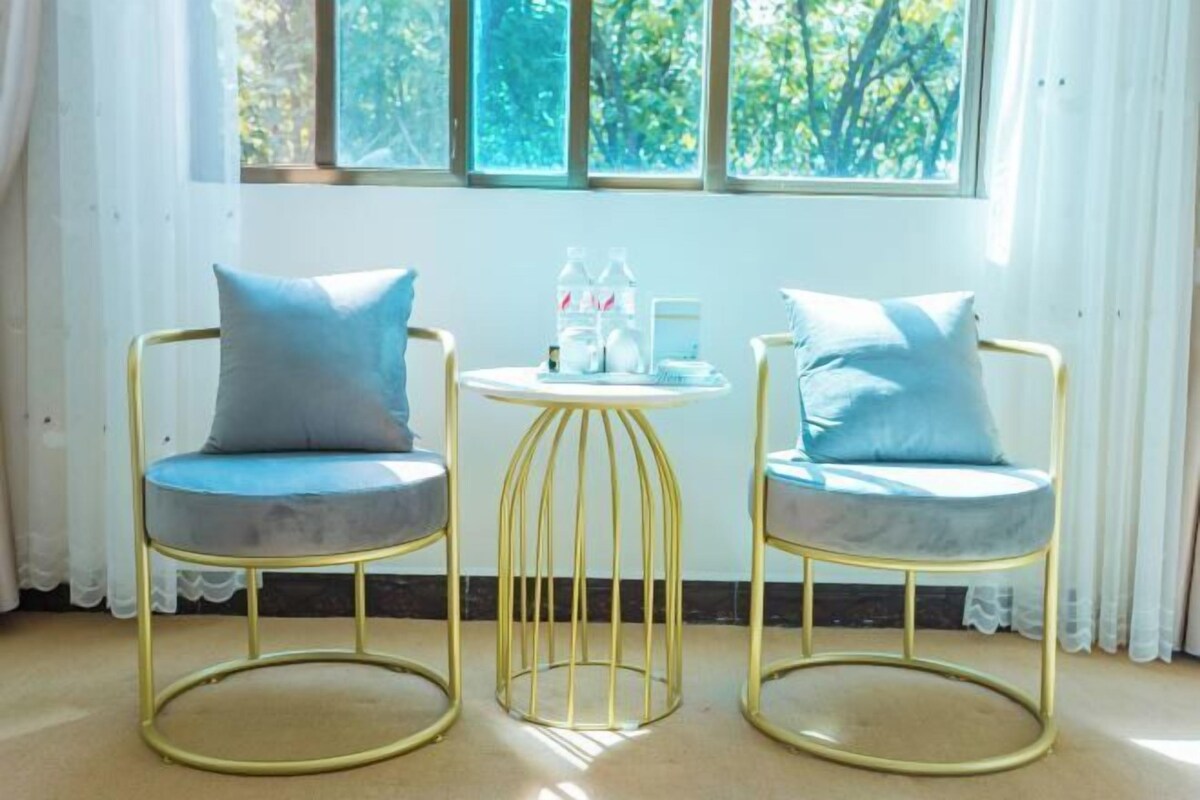 Two gold-framed chairs with blue cushions and a small round table with a white base, positioned in front of a window with white curtains.