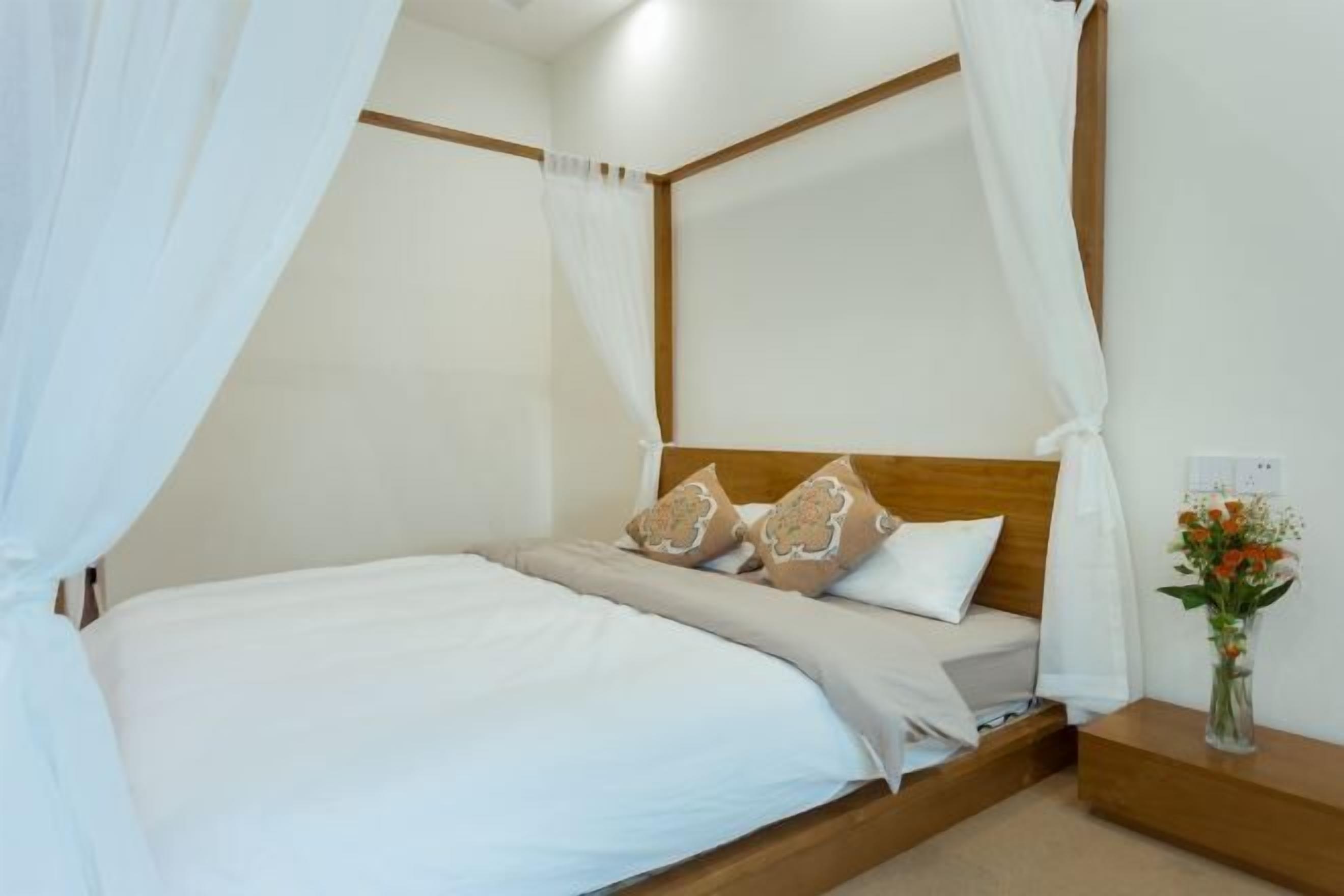 Una cama bien hecha con sábanas blancas y almohadones decorativos, un cabecero de madera y una mesita de noche con un jarrón de flores.