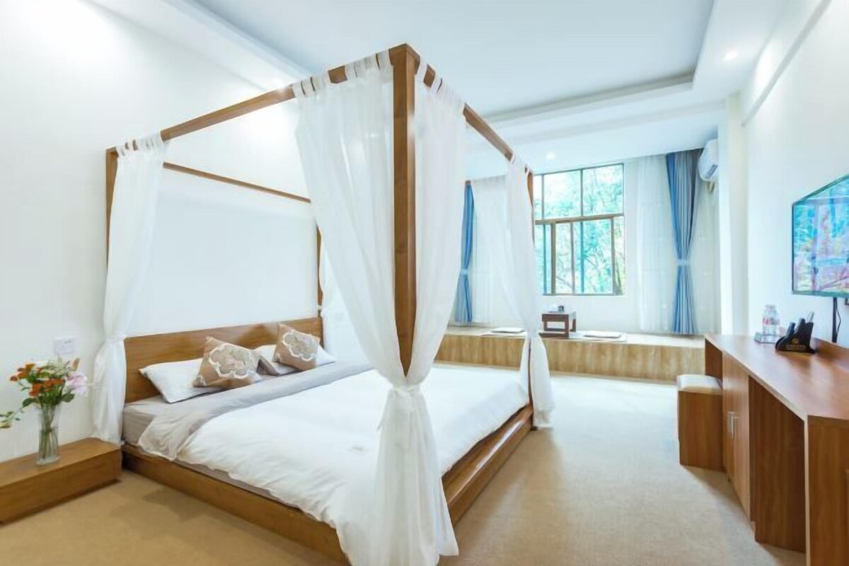 A bedroom with a four-poster bed, a bedside table, a desk, and a window with curtains.