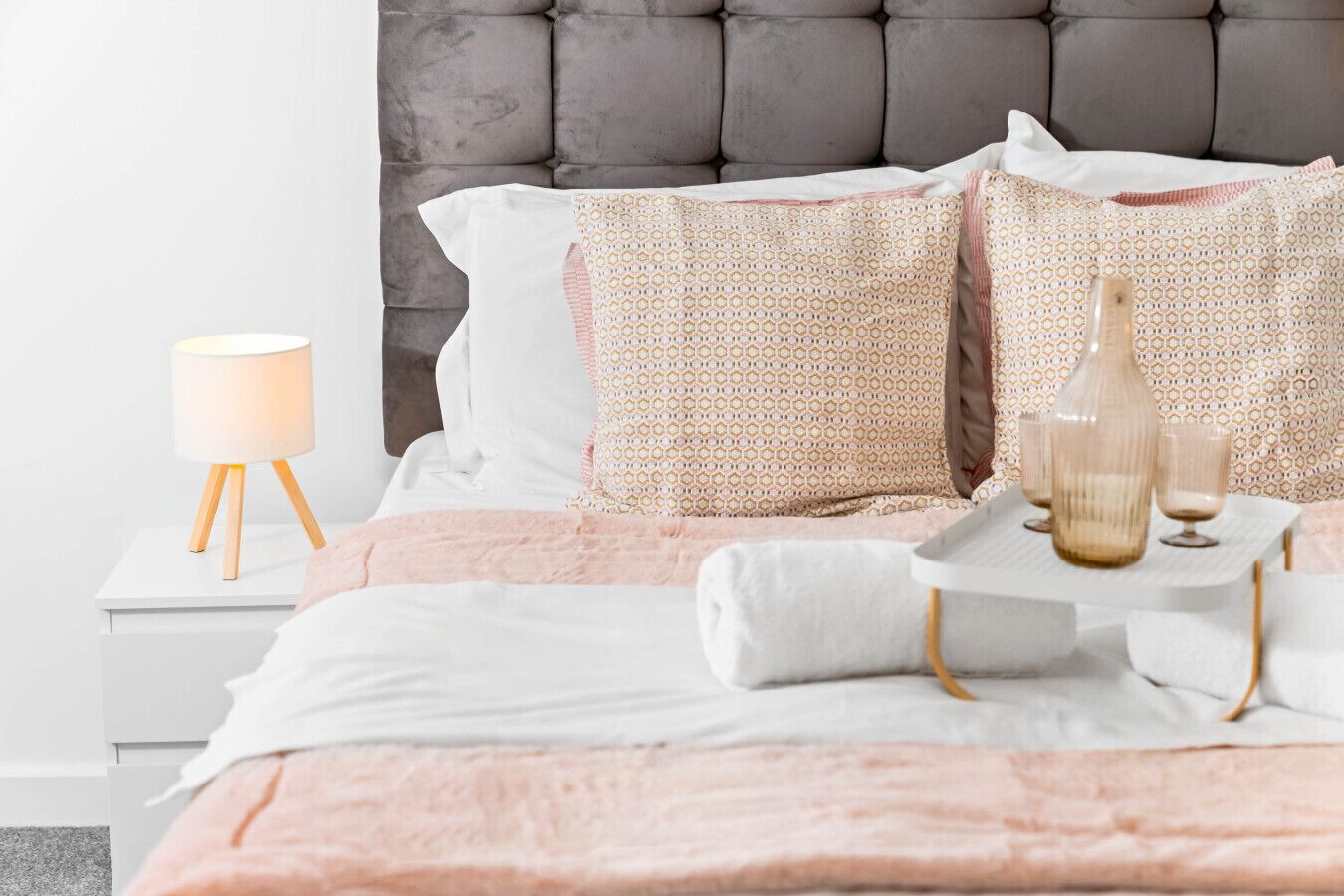 Un lit bien fait, recouvert d’une couverture rose, avec deux coussins à motifs, une lampe de chevet et un plateau contenant des verres et une bouteille.