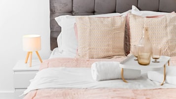 Un lit bien fait, recouvert d’une couverture rose, avec deux coussins à motifs, une lampe de chevet et un plateau contenant des verres et une bouteille.