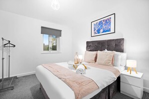 A modern bedroom with a large bed, a bedside table with a lamp, a window with a view, and a hanging coat rack. - Luxury Two Bedroom Apartment - Parking - Smart TV (Birmingham)