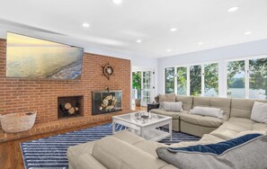 Un salon moderne avec une cheminée en briques, un grand téléviseur à écran plat, un canapé d’angle beige et une vue sur un lac, visible à travers de grandes fenêtres.