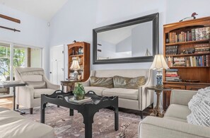 A living room with a sofa, armchairs, a coffee table, a bookshelf, a mirror, and a lamp.