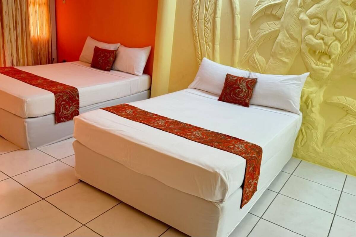 Dos camas individuales con ropa blanca y detalles decorativos rojos, piso de baldosas y una pared con cabecera tallada.