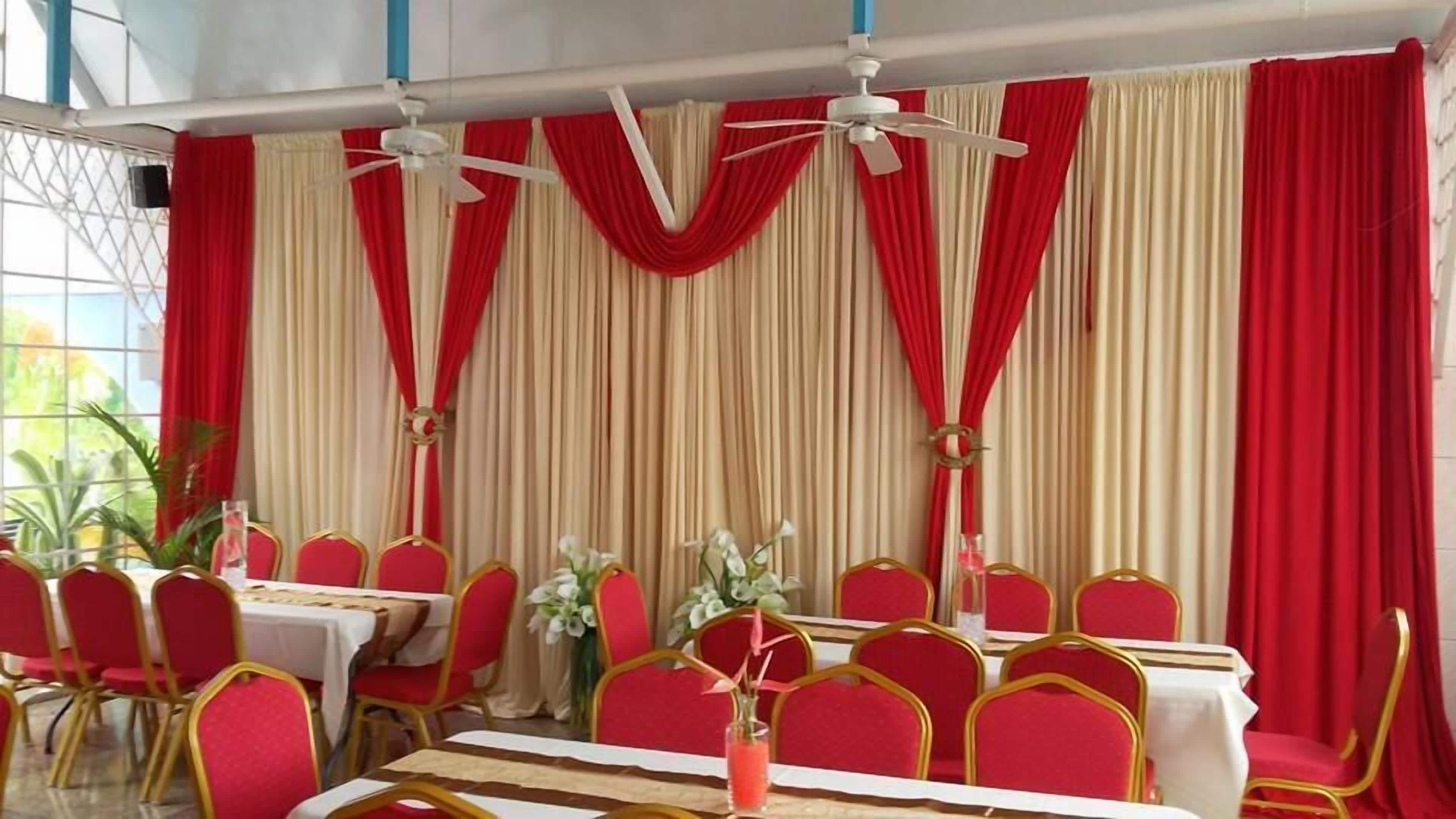 Un salón de banquetes con cortinas rojas y crema, ventiladores de techo blancos y sillas rojas con detalles dorados.