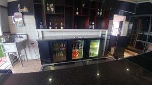 A bar area with a stainless steel sink, a refrigerator with multiple shelves, and a variety of bottles and glasses. - Alicia's Palace (Port of Spain)