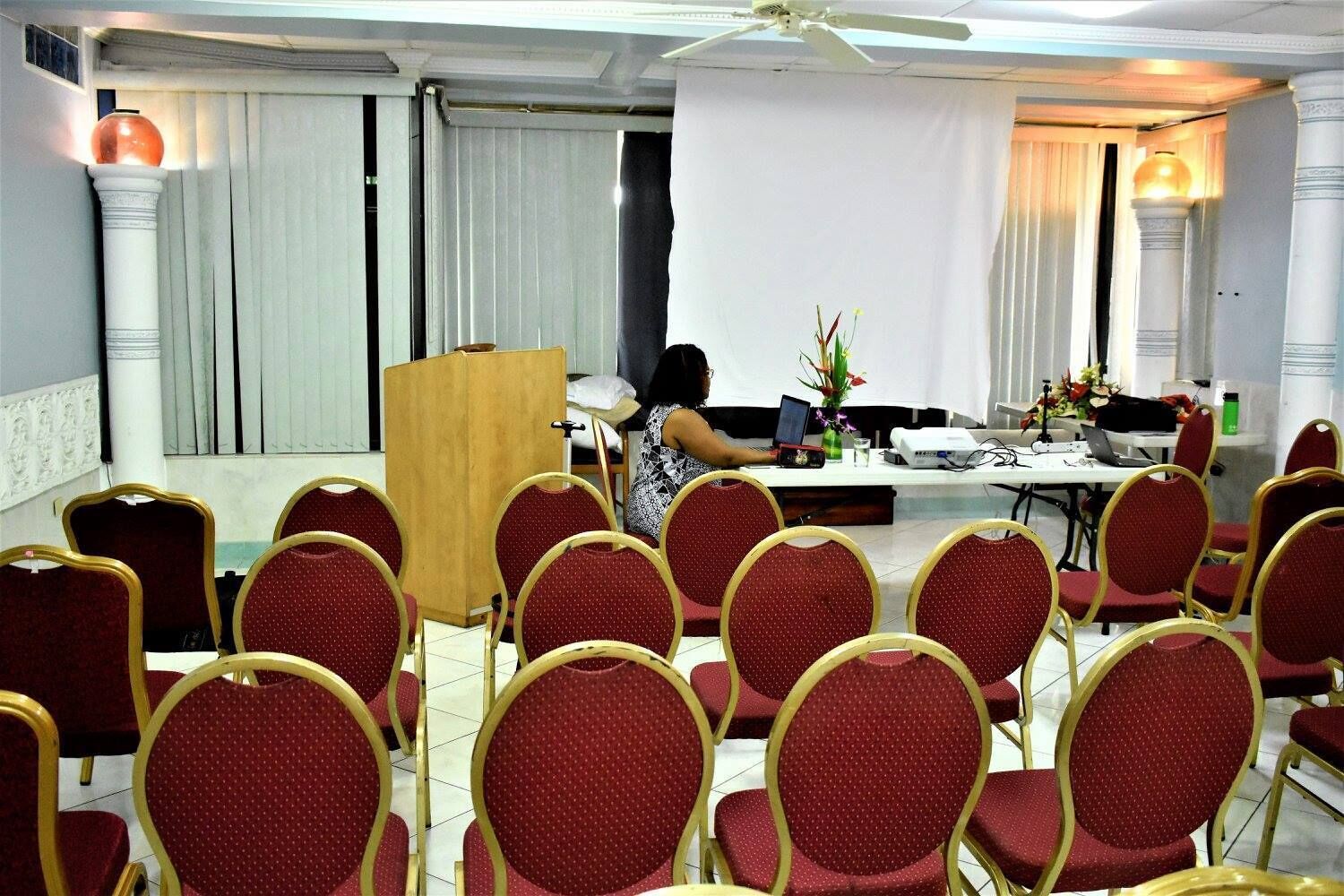Un salón de conferencias con filas de sillas rojas, un atril y una mesa con una computadora portátil y flores.