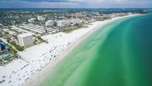 An aerial view of a coastal area with a long sandy beach, a clear blue ocean, and a dense cluster of buildings in the background. - Sarasota Home Away From Home (Sarasota)