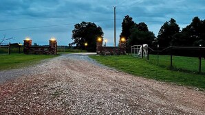 Uma estrada de cascalho leva a um portão de pedra com luzes, cercado por grama e árvores.