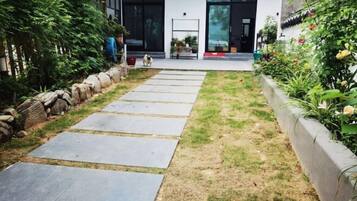 Een pad dat leidt naar een modern gebouw met een glazen deur, omgeven door een tuin met een stenen rand en verschillende planten.