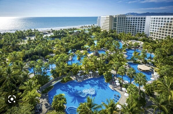 Ein Resort mit mehreren Schwimmbecken, umgeben von Palmen, einem Strand und einem großen Hotelgebäude.