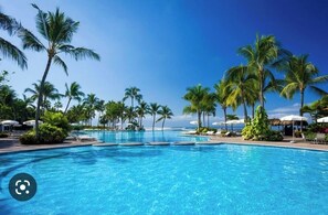 Ein großer Pool, umgeben von Palmen und Sonnenschirmen, mit blauem Himmel im Hintergrund.