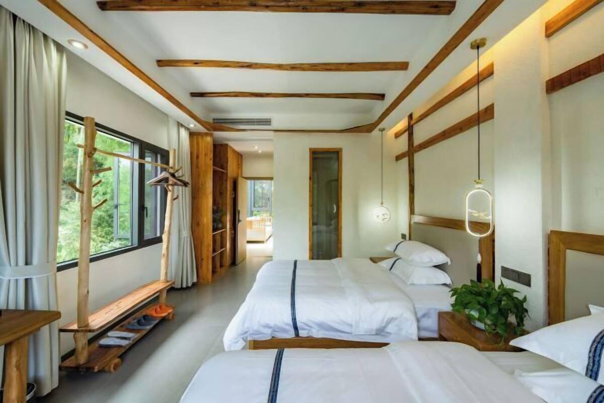 A room with three beds, wooden beams on the ceiling, a large window with curtains, and a plant on the bedside table.