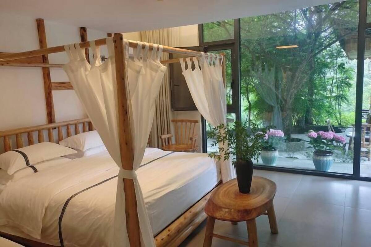 A room with a four-poster bed, a wooden headboard, a small wooden stool, and a large window with a view of greenery.