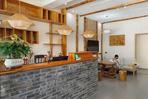 Una zona de recepción con un mostrador de madera, una planta grande en maceta y un área de estar con sofá y una mesita.
