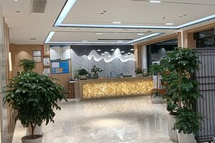 A hotel reception area with a modern design, including a reception desk, potted plants, and a wall-mounted information board.