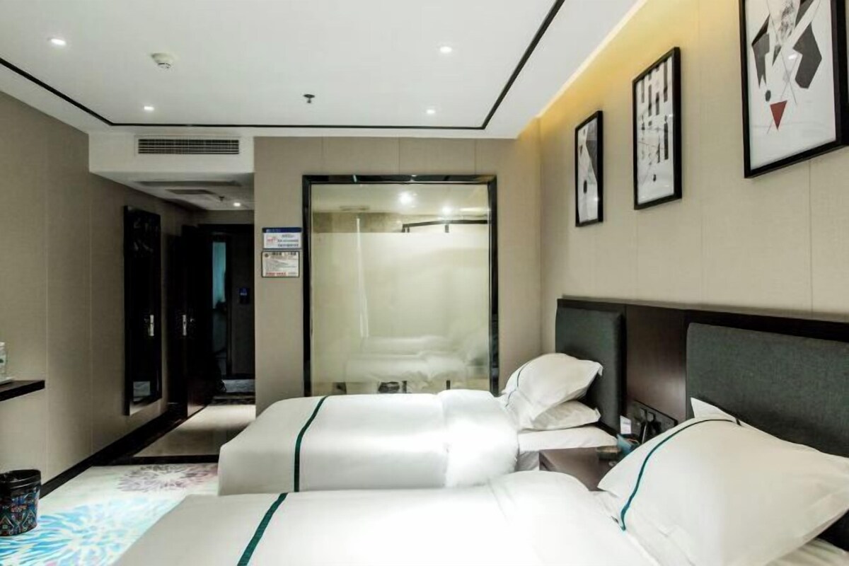Habitación de hotel con dos camas, baño con puerta de vidrio y cuadros en la pared.