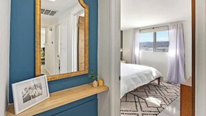 Habitación de hotel con un espejo grande, una mesita de noche con un marco de fotos, una cama con colcha blanca y vistas a la ciudad desde la ventana.