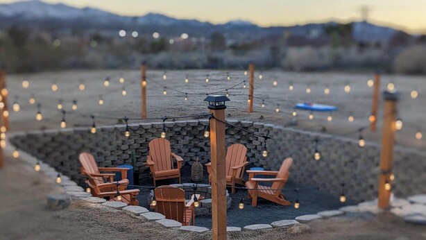 Un área de estar al aire libre con sillas y mesas de madera, rodeada de muros de piedra y luces colgantes.