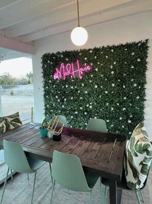 Un'area pranzo con tavolo in legno, sedie verdi e un neon su una parete ricca di verde.