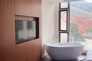 Un baño moderno con una bañera independiente, un televisor montado y un ventanal con vistas a una obra en construcción.