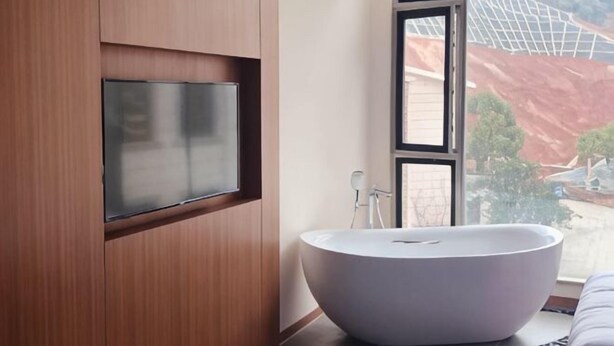 Un baño moderno con una bañera independiente, un televisor montado y un amplio ventanal con vistas a una obra en construcción.