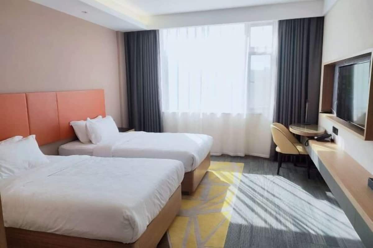 Habitación de hotel con dos camas, un escritorio, una silla, un televisor y ventanas amplias con cortinas.
