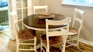 Un coin repas avec une table ronde et quatre chaises blanches à assise en osier, un plancher en bois, une porte blanche et une fenêtre avec des stores.