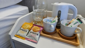 Una bandeja con cafetera, un vaso de agua, dos tazas de café y dos porciones de bocadillos sobre una mesita de noche blanca.