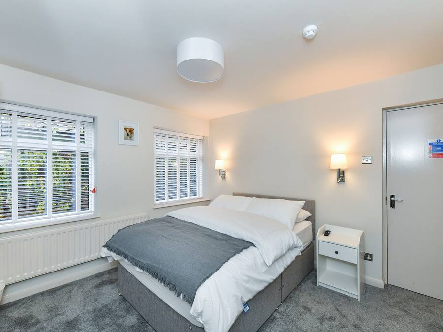 A bedroom with a bed, bedside tables, a TV, and two windows with blinds.