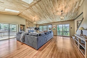 Ein großzügiger Wohnbereich mit einer großen grauen Couch, Holzboden, einem Steinkamin und großen Fenstern mit Blick ins Freie.