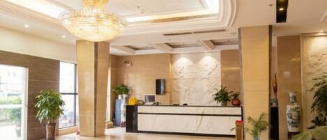 A hotel lobby with a reception desk, large chandelier, and potted plants.