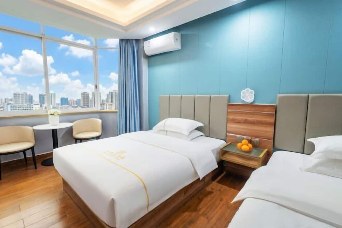 Habitación de hotel con un ventanal grande, una cama, una mesita de noche con fruta y vistas a la ciudad.
