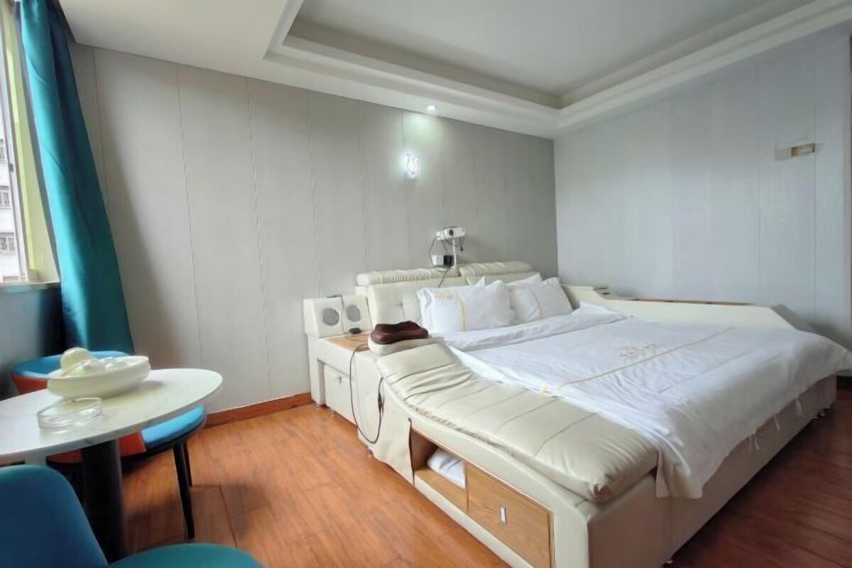 A hotel room with a bed, a small table with a chair, a bedside table with a lamp, and a window with curtains.