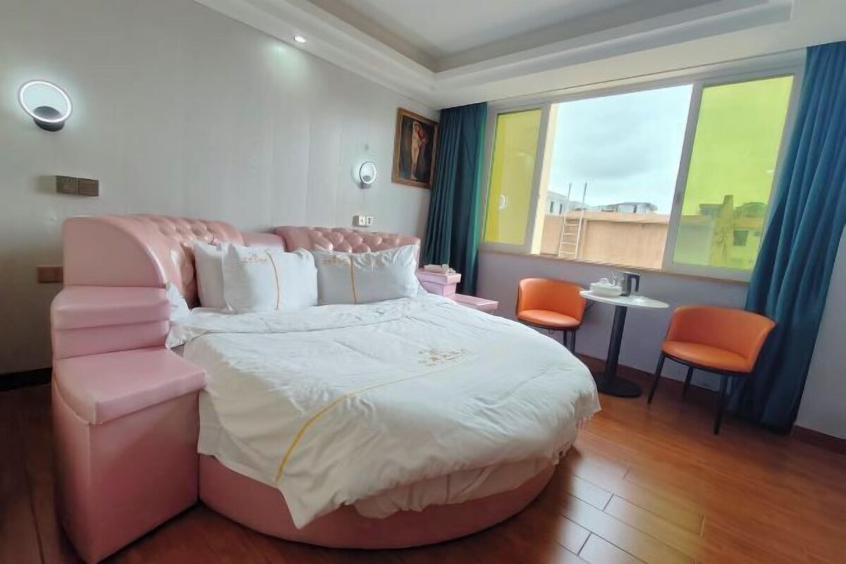 A modern bedroom with a round bed, a small table, two chairs, and a large window with curtains.