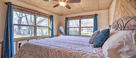 A bedroom with a ceiling fan, large windows with curtains, a bed with patterned bedding, and a view of the outdoors.
